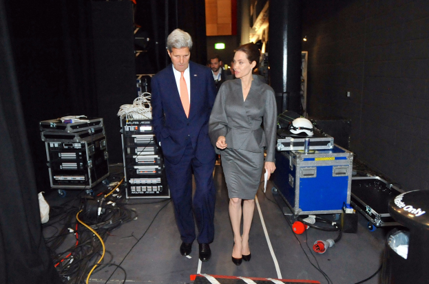 Angelina Jolie und John Kerry auf einem Boden umgeben von Menschen, Geräten, Kabeln, einem Helm auf einem Wagen, einem Müllcontainer, einem Vorhang, einer Säule und einer Decke mit Lampen.