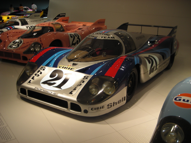 Ein Porsche 917K Le Mans-Rennwagen in einem Museum ausgestellt, umgeben von anderen Sportwagen, mit Menschen in der Nähe und einer Wand im Hintergrund.