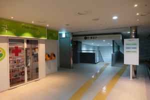 Apothekeneingang mit einem "Apotheke"-Schild auf der linken Seite, einer Tafel mit Text auf der rechten Seite, Hintergrundobjekte und beleuchteter Dachvorsprung.