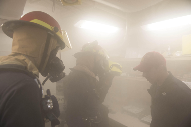 Feuerwehrleute mit Helmen und Rauch aus dem Mund, einer hält eine Kamera, während einer Übung mit Licht und Equipment im Hintergrund.