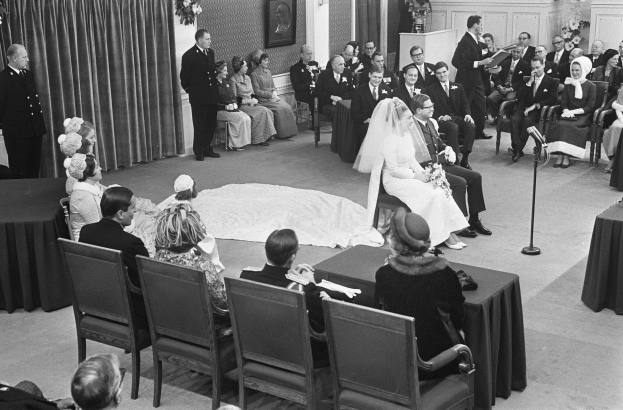 Schwarze und weiße Fotografie einer Hochzeitszeremonie mit sitzenden und stehenden Gästen, einem Mann mit Mikrofon, einer Frau mit Blumenstrauß, einem Podium mit Mikrofon, Blumenschmuck, einem Vorhang, einem gerahmten Foto an der Wand und einer Tür im Hintergrund.