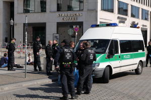 Eine Gruppe von Polizisten steht vor einem Starbucks-Café, rechts davon parkt ein Van und links stehen und sitzen ein paar Menschen; im Hintergrund sind ein Gebäude, ein Schild, ein Laternenpfahl und ein Zaun zu sehen.