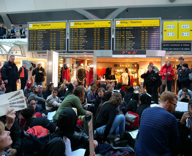 Eine große Gruppe von Menschen an einem Flughafen, einige mit Taschen und Papieren sitzend, während andere stehen, mit Texttafeln, Schaufensterpuppen in Kleidern und Deckenleuchten im Hintergrund, was auf eine Protestveranstaltung hindeutet.