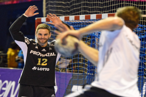 Zwei Männer spielen Handball vor einem Tor mit Netz und einem Banner mit Text im Hintergrund und einigen Zuschauern in einer Arena.
