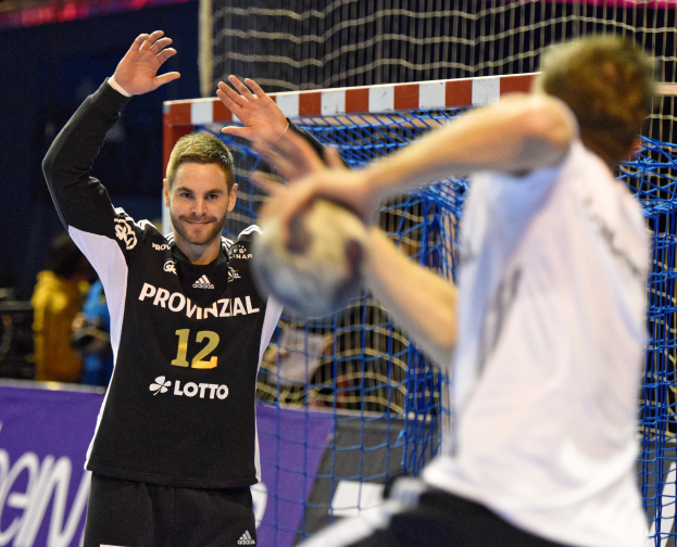 Zwei Männer spielen Handball vor einem Tor mit Netz und einem Banner mit Text im Hintergrund und einigen Zuschauern in einer Arena.