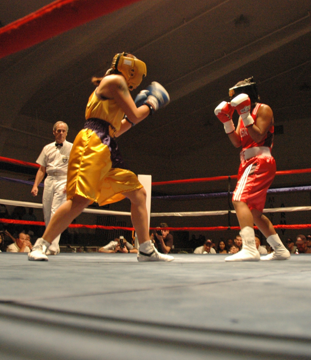 Zwei Boxer im Ring mit einem Schiedsrichter und Zuschauern, einige Fotografen, unter einer sichtbaren Decke.