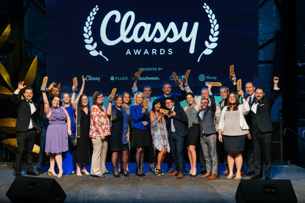 Gruppe von Menschen auf einer Bühne, die Auszeichnungen halten, mit Lautsprechern darunter und einem Bildschirm im Hintergrund mit der Aufschrift "Classy Awards 2019"