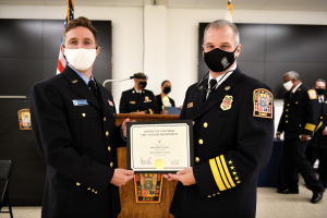 Zwei Männer in Uniform und Masken stehen nebeneinander und halten ein Zertifikat, mit einem Podium mit Mikrofon dahinter und einem Stuhl links, in einem Raum mit Menschen, Flaggen und einer Wand im Hintergrund.