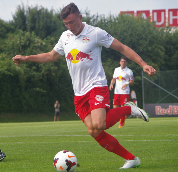 Ein Mann in einem Fußballtrikot schießt einen Ball auf einem grünen Feld mit Bäumen und einem klaren blauen Himmel im Hintergrund, wobei ein Schild mit der Aufschrift "RB Leipzig" zu sehen ist.