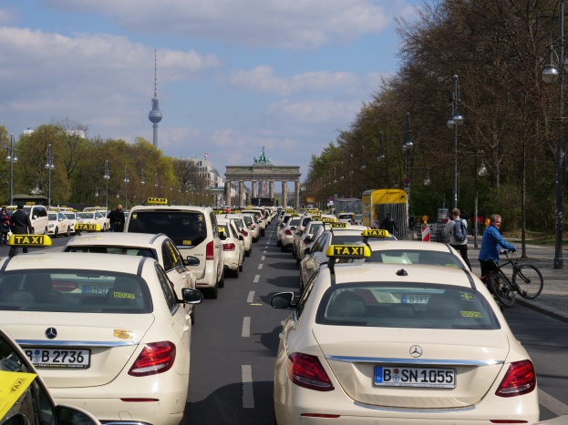 Eine lange Reihe von Taxis, die auf der Seite einer belebten Straße in Berlin, Deutschland, geparkt sind, mit Fahrzeugen, Radfahrern und Fußgängern, flankiert von Laternenmasten, Bäumen und Gebäuden, einschließlich eines Bogens und eines Turms, unter einem bewölkten Himmel.