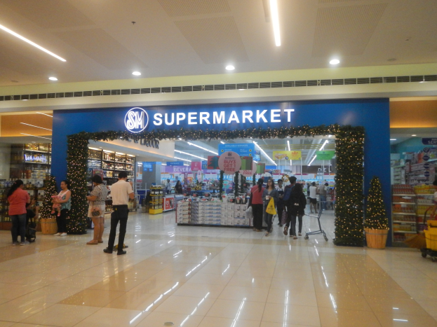 Gruppe von Menschen vor einem Supermarkt mit Einkaufstägern, einem Wagen, Topfpflanze, Regalen mit Gegenständen, Schildern und einer Decke mit Deckenleuchten.