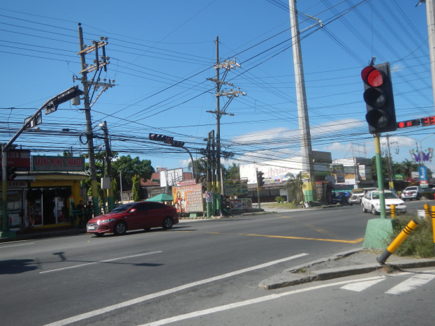 Ein rotes Auto fährt an einer Ampel mit Strommasten und Leitungen entlang der Straße vorbei, Gebäude und Bäume im Hintergrund unter einem klaren blauen Himmel.