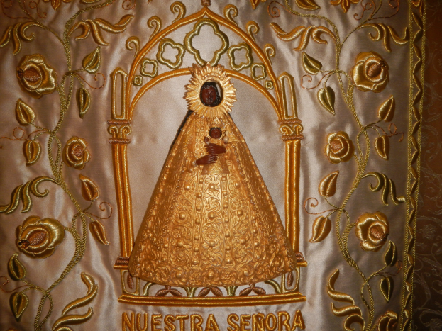 Goldbestickte Chasuble mit einer serenitätsstrahlenden Frau in einem hochgeschlossenen, langärmeligen Kleid mit einem Dutt, umgeben von kunstvollen Designs und prunkvollen Texten in einem goldenen Rahmen.