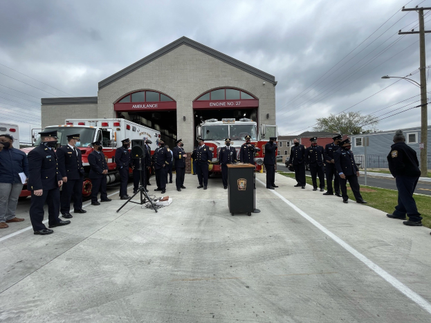 Gruppe von Menschen in Uniformen neben einem Löschfahrzeug auf einem Podium bei einer Feuerwache-Einweihung, mit Gebäuden, Bäumen, Strommasten und Gras im Hintergrund unter einem bewölkten Himmel.