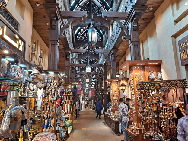 Ein geschäftiges Dubai-Bazar mit Menschen, die zwischen Regalen voller Gegenstände hindurchgehen, Wänden mit Text und einer Decke mit warmem Licht.