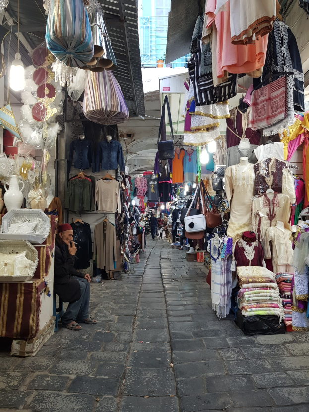 Ein belebter Straßenmarkt in der Altstadt von Jerusalem mit Menschen, die an Ständen mit Kleidung, Taschen, Lampen und anderen Artikeln vorbeischlendern, und einem Gebäude im Hintergrund.