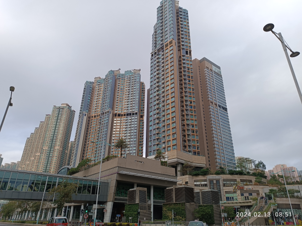 Eine Stadtstraße mit hohen Gebäuden, Fahrzeugen, Laternen, einer Brücke, Bäumen und einem bewölkten Himmel, mit einem prominenten Wohnhaus im Vordergrund.