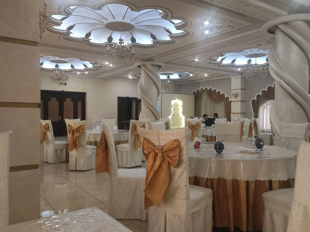 Ein großer Festsaal mit Tischen und Stühlen, die mit Tüchern bedeckt und mit verschiedenen Gegenständen geschmückt sind, mit Türen und Vorhängen an den Wänden und beleuchtet von Deckenlampen und Kronleuchtern.