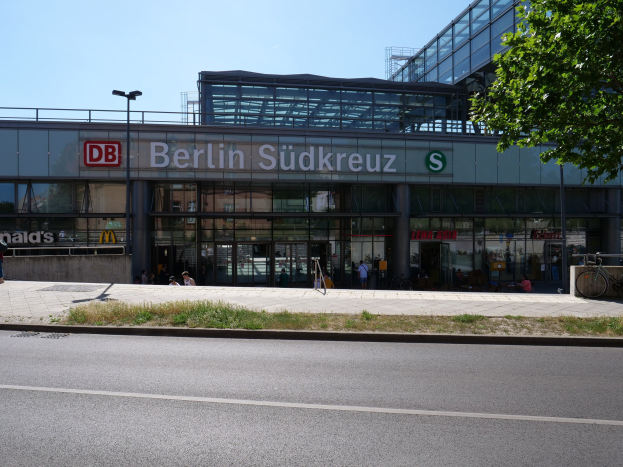 Großes Glaswand-Bahnhof namens "Südkreuz" in Berlin, Deutschland, mit Straßenlaternen, Lichtern, Fahrzeugen, Fußgängern, Fahrrädern, Bäumen und einem klaren blauen Himmel.