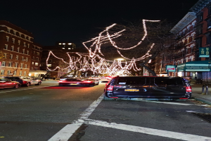 Eine nächtliche Straßenansicht mit parkenden Autos, Gebäuden mit Fenstern auf beiden Seiten, einem beleuchteten Baum im Hintergrund und einem Van im Vordergrund unter einem dunklen Himmel.