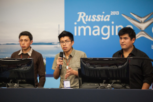 Drei Jungen stehen neben einem Schreibtisch mit zwei Computersystemen, einer spricht in ein Mikrofon und trägt eine ID-Karte mit roter Markierung, vor einer Wand mit einer Werbetafel, auf der 'Russia Imagine 2013' steht.