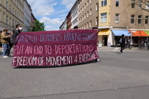 Eine Gruppe von Menschen marschiert auf einer Stadtstraße und hält ein Transparent mit der Aufschrift "Abolish Borders, Abolish Frontiers, Put an End to Deportations, Freedom of Movement 4 Everyone". Im Hintergrund sind Gebäude, Bäume, Fahrräder und ein bewölkter Himmel zu sehen.