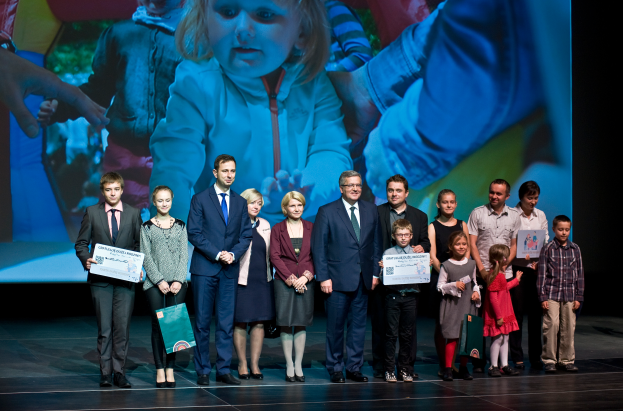Eine Gruppe von Menschen auf einer Bühne, einige halten Taschen und Urkunden, mit einem Bildschirm im Hintergrund, der ein Bild eines Kindes und anderer Menschen anzeigt.