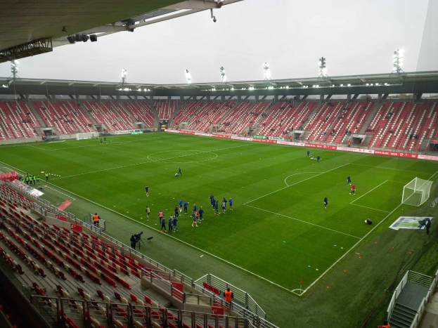 Ein Fußballfeld in einem Stadion mit Menschen, Sitzplätzen, Geländern, Stufen und Lichtern, mit dem Himmel im Hintergrund.