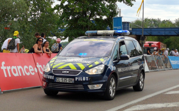 Polizeiauto fährt an einer Menge vorbei, die Schilder hält, eine Brücke, Bäume, eine Fahne und einen bewölkten Himmel im Hintergrund.