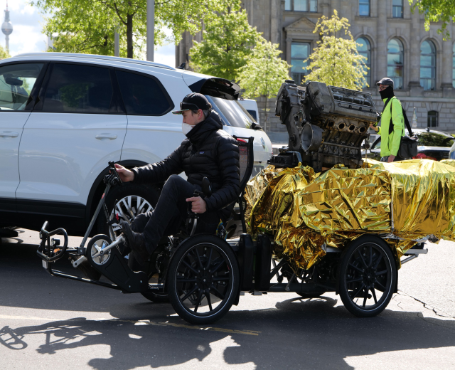 Ein Mann im Rollstuhl mit einem großen Motor auf seinem Rücken, umgeben von Fahrzeugen auf einer Straße mit Bäumen, Gebäuden, Polen und einem klaren blauen Himmel im Hintergrund; er trägt eine schwarze Jacke, eine Mütze und hält ein Objekt in der Hand.