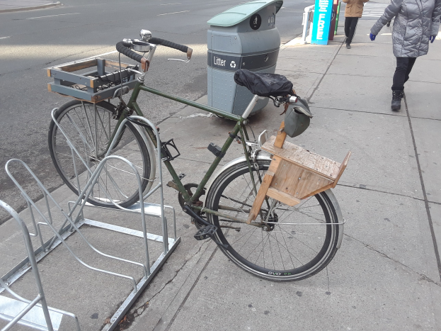 Ein grünes und braunes Fahrrad neben einem Müllcontainer auf einer Straße mit Fahrzeugen und Fußgängern im Hintergrund.