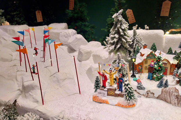 Eine festliche Weihnachtsdorf-Szene mit schneebedeckten Elementen, darunter ein Berg im Hintergrund, Tannen, Häuser, Menschen, Fahnen, Masten, Lichter und Tiere, alles unter einem dunklen Himmel und damit eine gemütliche Atmosphäre.