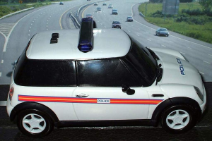 Ein weißer Polizeiwagen steht im Vordergrund mit mehreren Autos, die dahinter auf der Straße fahren, umgeben von Gras und Bäumen auf beiden Seiten.