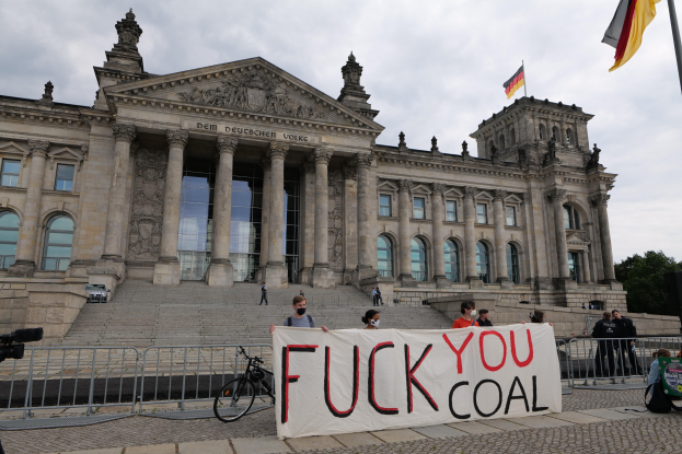 Eine Gruppe von Menschen mit einem Transparent "Fuck You Coal" vor dem Reichstaggebäude in Berlin, Deutschland, mit Bäumen und einer Fahnenstange im Hintergrund.