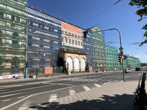 Großer mehrgeschossiger Bau unter construction in Berlin, umgeben von Straßeninfrastruktur, Fahrzeugen, Fußgängern und Bäumen unter einem bewölkten Himmel.