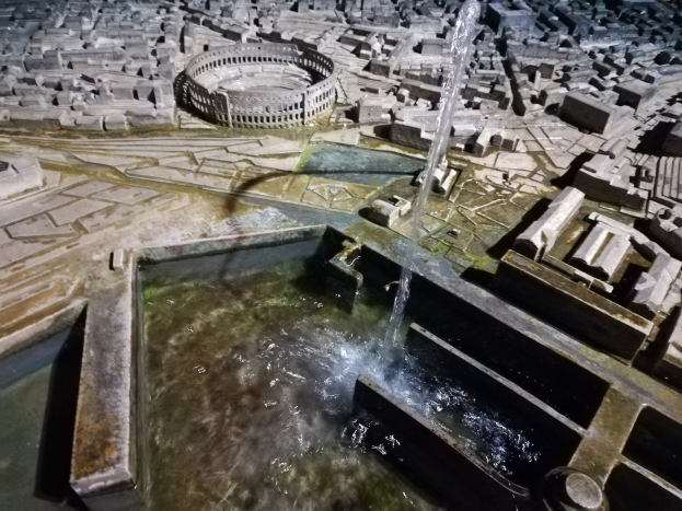 Ein Miniaturstadtmodell mit einem zentralen Brunnen mit Wasserfall, umgeben von Gebäuden und Wasserflächen.