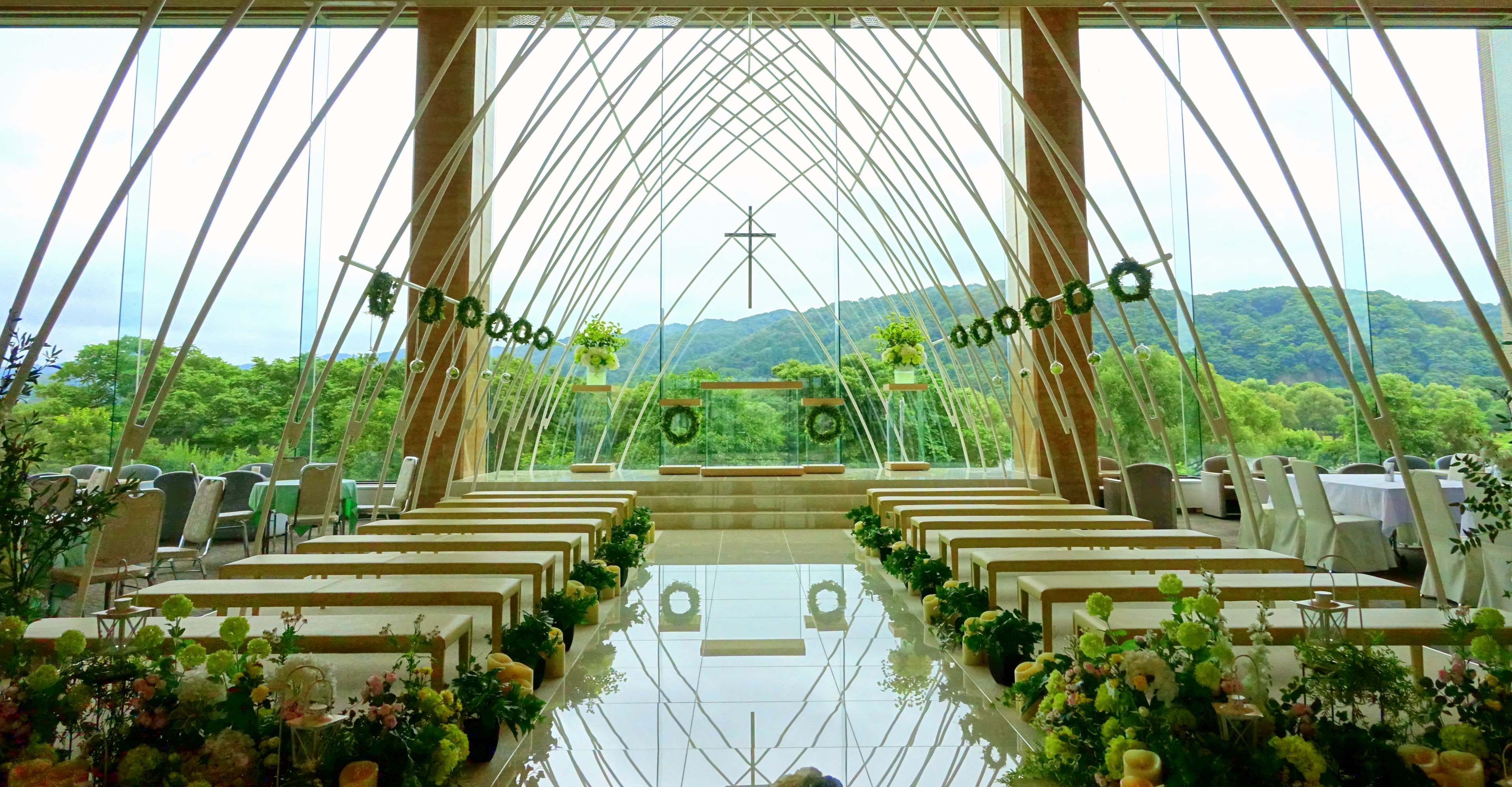 Eine Hochzeitszeremonie in einem großen, geschmückten Saal mit halbrunder Bestuhlung, Blumenschmuck und einer Aussicht auf Bäume, Hügel und einen blauen Himmel im Hintergrund.