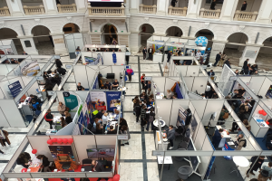 Große Besucherzahl auf einer internationalen Fachmesse mit Ständen, Bannern, Tischen, Stühlen und Ballons in einem Raum mit architektonischen Bögen und Säulen.