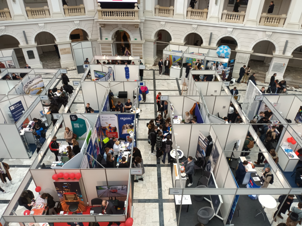 Große Besucherzahl auf einer internationalen Fachmesse mit Ständen, Bannern, Tischen, Stühlen und Ballons in einem Raum mit architektonischen Bögen und Säulen.