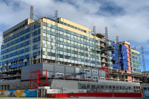 Großes Gebäude im Bau in einer städtischen Umgebung mit Glasfenstern, Metallrahmen, einer Treppe, einem Zaun, einem Schild, Polen, einem Straßenpfahl, umliegenden Bäumen und einem bewölkten Himmel.