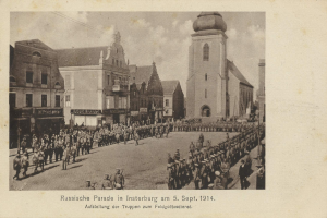 Ein Schwarz-Weiß-Foto eines Umzugs in Insterburg am 5. September 1914 mit vielen Menschen, Gebäuden und bewölktem Himmel sowie einem Text am unteren Rand.