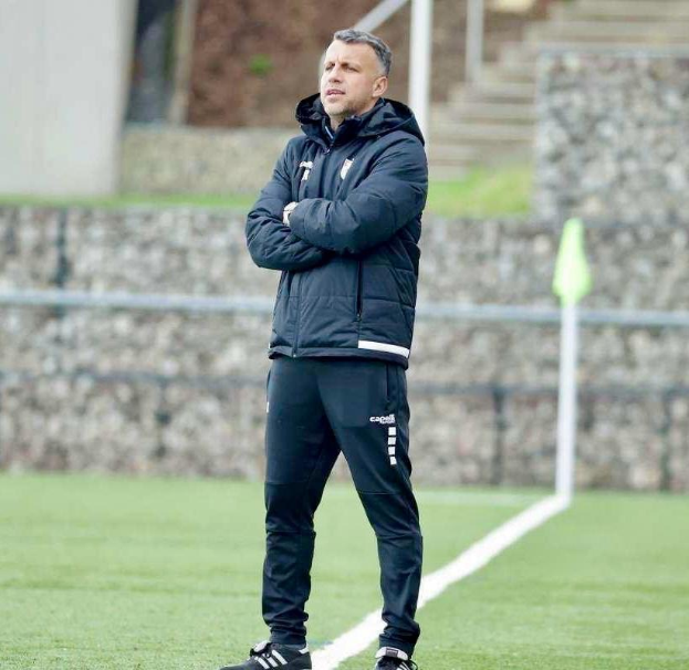 Ein Fussballtrainer, der auf einem Feld steht und die Ärmel überkreuzt, trägt eine schwarze Jacke und Hose, mit einem Zaun, Treppe und Wand im Hintergrund.
