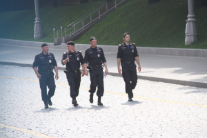 Drei Polizisten in Uniform und Mützen gehen eine Straße mit Pfählen, Geländern, Treppen und Gras im Hintergrund entlang.
