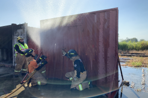 Drei Feuerwehrleute in Helmen und Jacken, einer spritzt Wasser aus einer Leitung auf einen Container, mit Bäumen, Pflanzen und einem klaren blauen Himmel im Hintergrund.