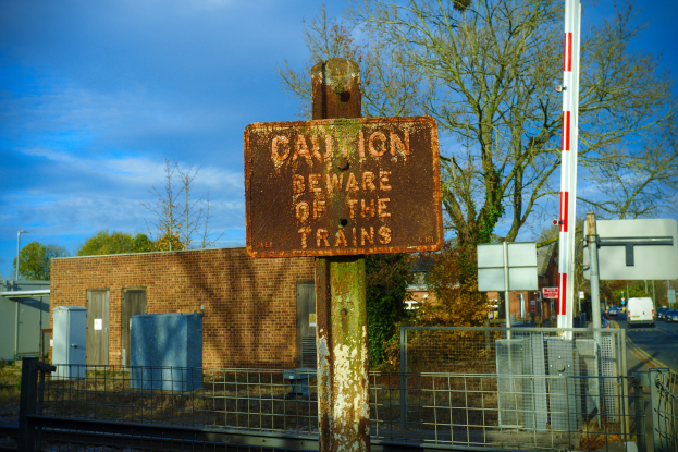 Vorsichtsschild an einer Bahnschiene neben einem Metallzaun, mit Bäumen, Strommasten, einem Gebäude, Containern, Fahrzeugen auf einer Straße und einem bewölkten Himmel.