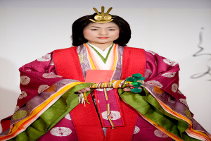 Eine Frau in einem traditionellen japanischen Kimono, das ein leuchtendes rotes und grünes Kleid mit aufwändigen Mustern trägt, steht selbstbewusst auf einem Laufsteg, ihr Haar ist ordentlich zu einem Dutt gebunden.