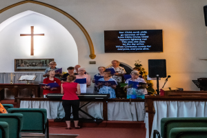 Eine Gruppe von Menschen steht vor einer Kirche, einige halten Bücher in den Händen, Stühle sind zu sehen, ein Kreuzsymbol und ein Photo Rahmen an der Wand, eine Tafel mit Text, ein Lautsprecher mit Ständer und ein Tisch mit einer Decke, wobei sie scheinbar als Chor singen.