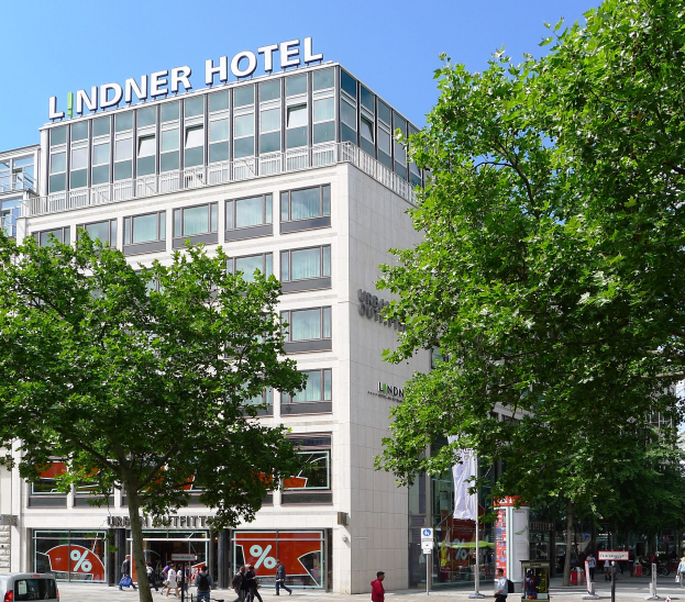 Großes Glasfrontgebäude mit der Aufschrift "Lindner Hotel" in Berlin, umgeben von Bäumen, Fahrzeugen, Passanten und Himmel.