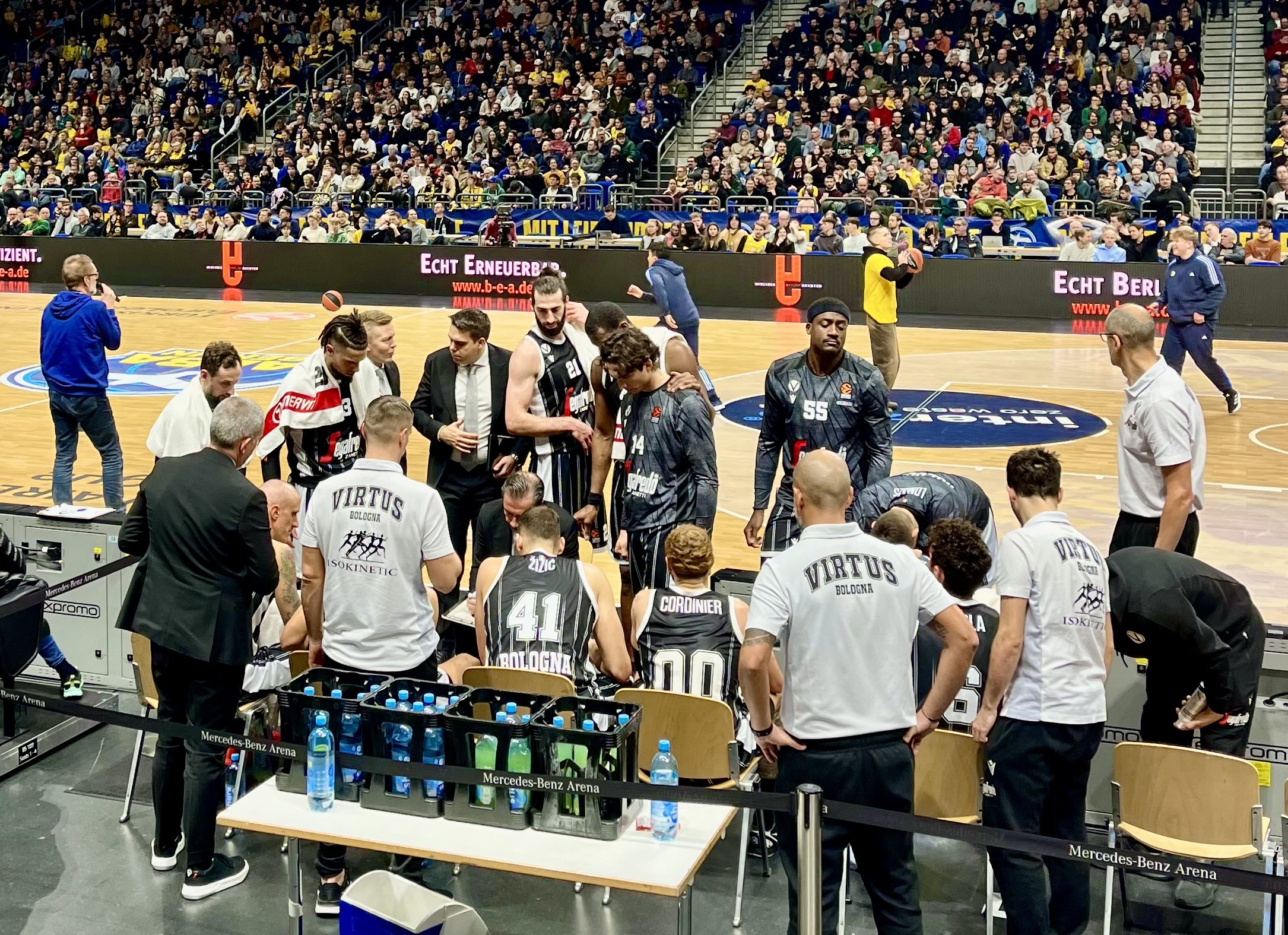 Eine Gruppe von Männern steht auf einem Basketballfeld während eines Spiels zwischen Virtus Berlin und Echt Berlin, umgeben von sitzenden und stehenden Zuschauern, mit einem Fotografen und Equipment links und Informationsschildern im Hintergrund.