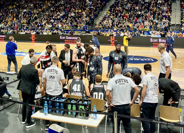 Eine Gruppe von Männern steht auf einem Basketballfeld während eines Spiels zwischen Virtus Berlin und Echt Berlin, umgeben von sitzenden und stehenden Zuschauern, mit einem Fotografen und Equipment links und Informationsschildern im Hintergrund.